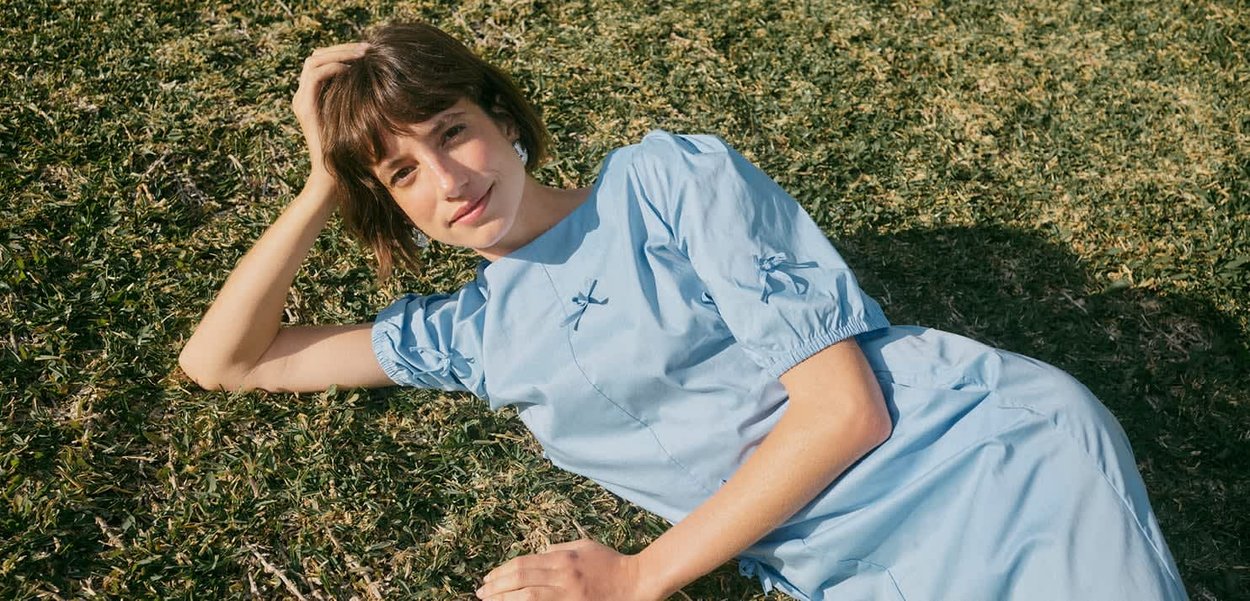 Junge Frau mit kurzen braunen Haaren, die ein hellblaues Kleid trägt, liegt auf dem Gras, stützt ihren Kopf auf die Hand und schaut in die Kamera.