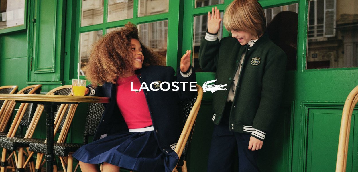 Due bambini che indossano abbigliamento Lacoste. Uno ha i capelli ricci, una camicia rossa e una gonna a pieghe blu scuro. L'altro indossa una giacca verde e pantaloni scuri. Sfondo verde.
