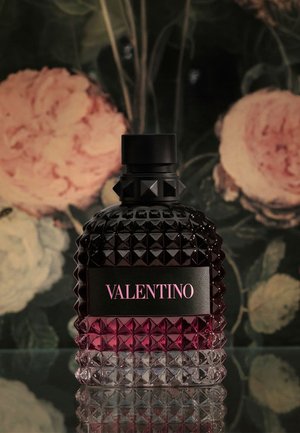 Dunkle strukturierte Valentino-Parfümflasche mit Nietenmuster und pinkem Etikett, vor einem blumigen Hintergrund mit großen pinken und weißen Blumen platziert.