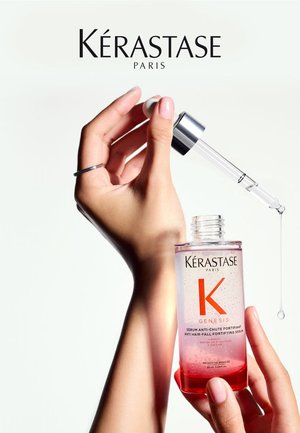 Siero anti-caduta dei capelli Kérastase Genesis in una bottiglia di vetro trasparente con un gradiente rosa a trasparente, contando su un contagocce argento e una goccia di siero visibile.