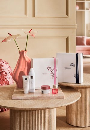 Vase rose avec deux arums, flacons de soin blanc, pot de crème, petit flacon de parfum rouge et deux boîtes cadeaux décoratives sur des tables en bois.