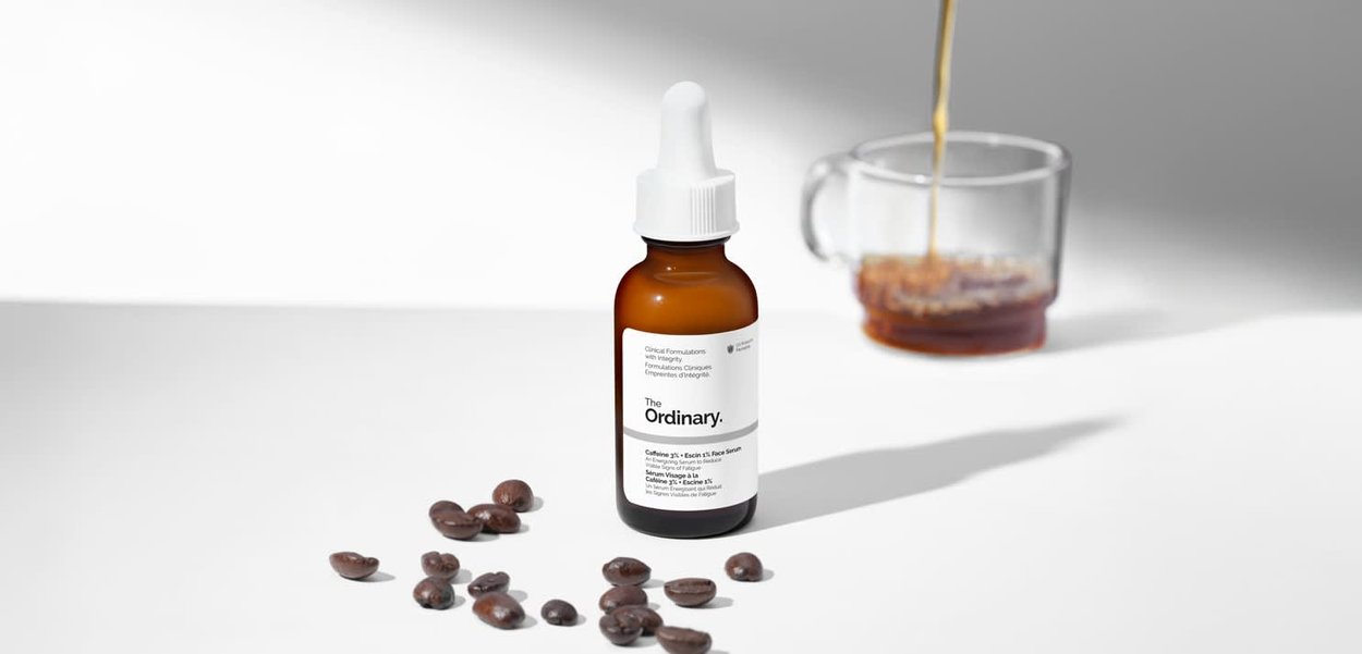 Botella gotero ámbar etiquetada como The Ordinary con granos de café esparcidos cerca y café vertiéndose en una taza de cristal en el fondo.