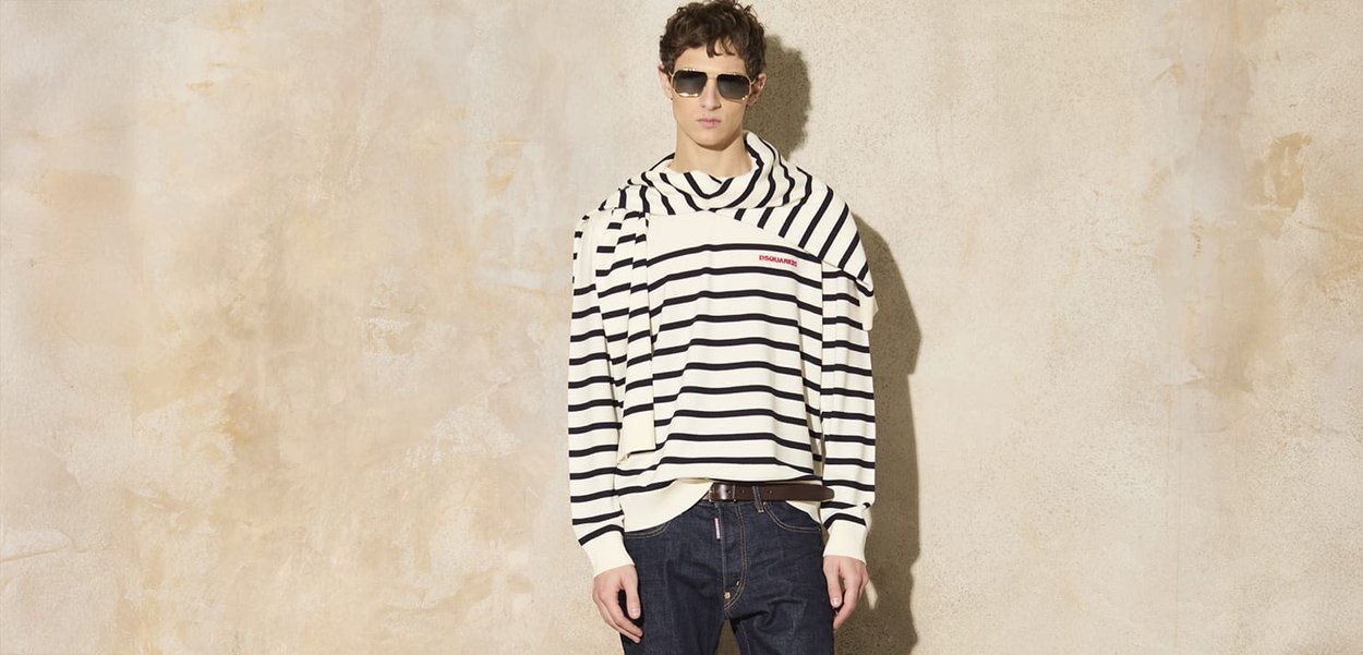 Pullover a righe beige e navy con un design drappeggiato unico; presenta un logo testuale ed è abbinato a jeans scuri. Il modello indossa occhiali da sole.