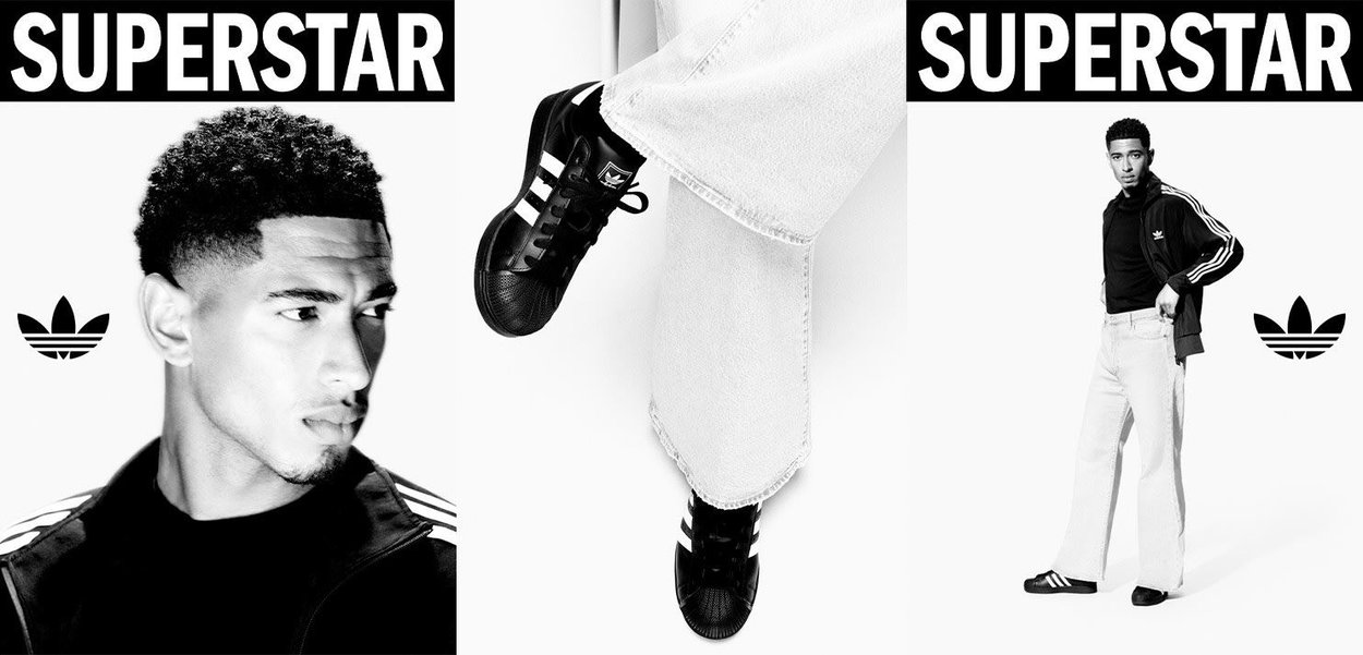 Zwarte Adidas Superstar sneakers met witte strepen, gecombineerd met een zwarte zip-up jas en lichte denim jeans. Dikke "SUPERSTAR" tekst erboven.