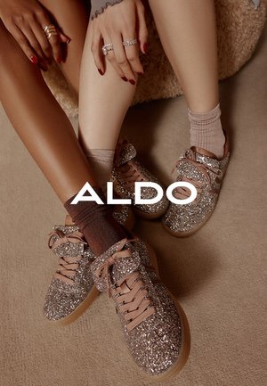 Glanzende zilveren sneakers met bruine veters en gum soles, gecombineerd met licht- en donkerbruine sokken, op een gestructureerd beige oppervlak.