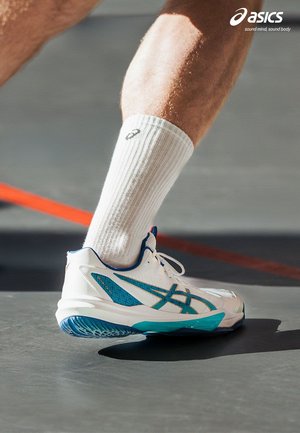 Idrettsutøverens underben iført hvit ASICS-sokk og hvit-blå ASICS idrettssko på innendørshall nær rød linje.
