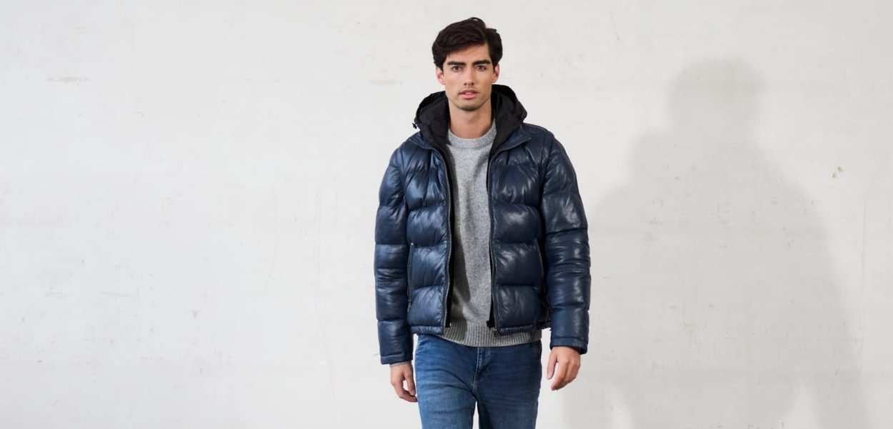 Giubbotto imbottito blu navy con zip frontale, cappuccio e design trapuntato. Indossato sopra un maglione grigio chiaro, abbinato a jeans blu. Texture liscia.