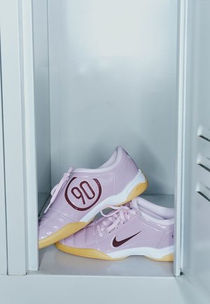 Nike-schoenen in lichtroze met een glanzende afwerking, uitgerust met een groot rood "90" logo, witte accenten en een rubberen zool, geplaatst in een kluis.