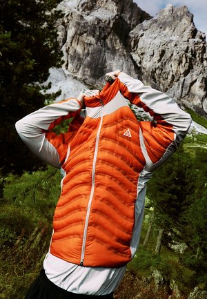 Persona che si infila una giacca arancione e grigia sopra la testa in un'area forestale montuosa con scogliere rocciose.