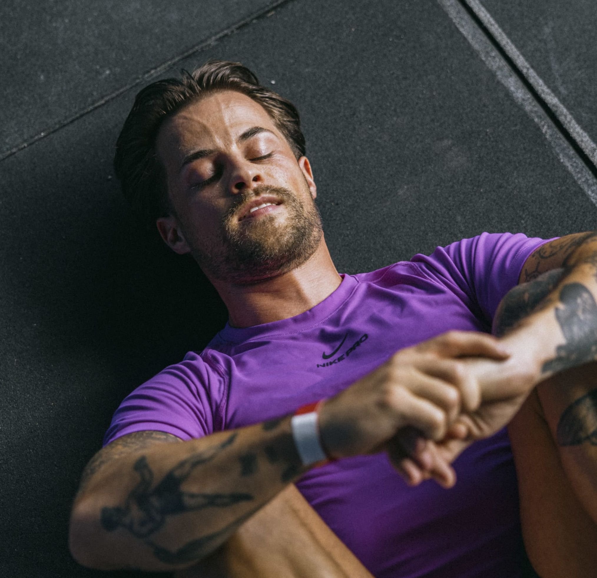 Purple Nike Pro athletic shirt with short sleeves, smooth texture. Person seated on a black surface, showing arm tattoos and a fitness wristband.