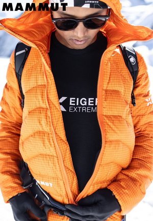 Person, der einen schwarzen Gürtel anpasst, während er eine orange Mammut-Jacke, schwarze Handschuhe, Sonnenbrille und ein schwarzes Eiger Extreme-Shirt im schneebedeckten Hintergrund trägt.