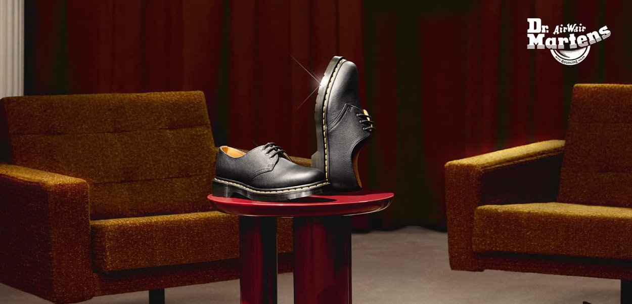 Scarpe Dr. Martens in pelle nera esposte su un tavolo rotondo rosso tra due poltrone imbottite marroni davanti a tende rosso scuro.