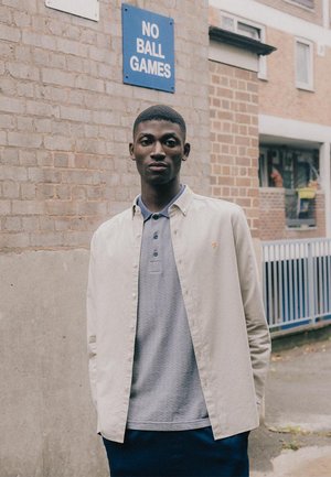 Chemise boutonnée beige clair sur un polo gris, avec un design texturé. Pantalons sombres, devant un mur en briques avec un panneau bleu.