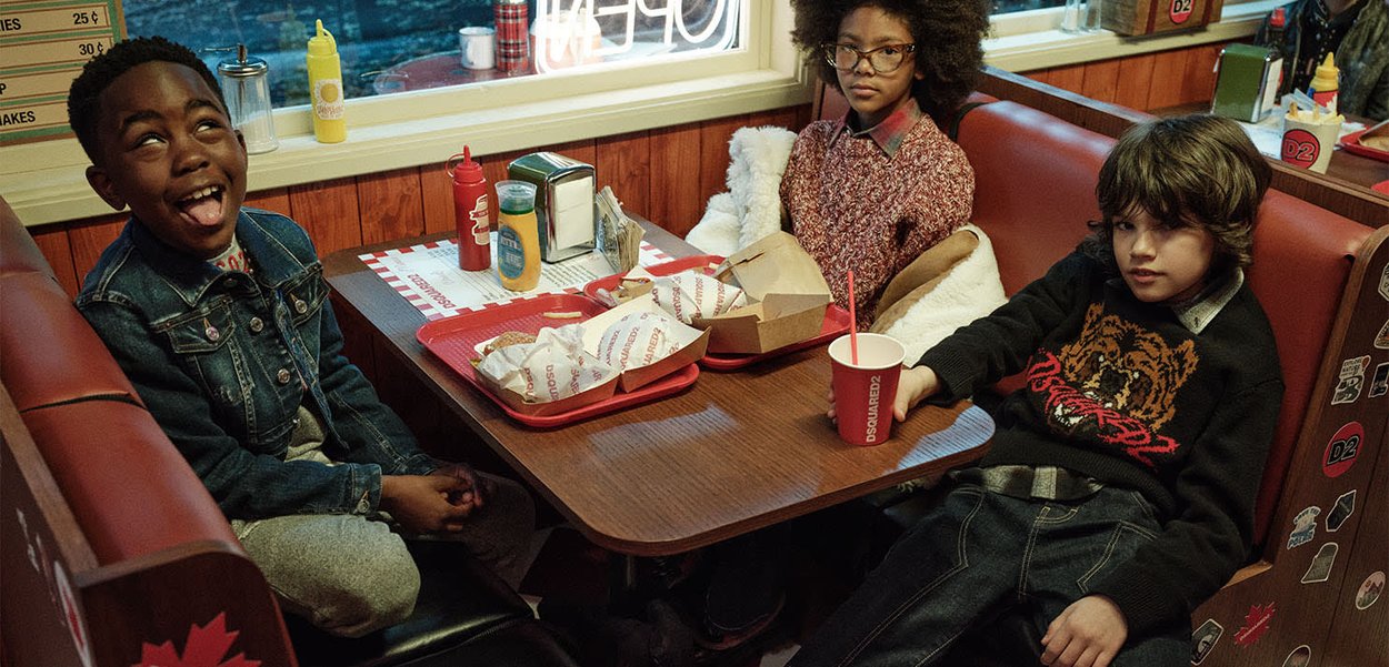 Drie kinderen zitten in een eetcafé in een booth met dienbladen vol ingepakte hamburgers, drankjes, sauzen en één kind steekt speels zijn tong uit.