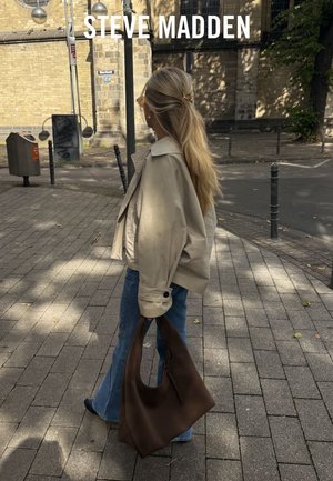 Beige korte jas, blauwe wijde jeans en een bruine slouchy handtas. Lichte achtergrond met een betegelde straat en minimale straatmeubilering.