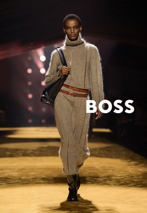 Modelka w beżowej dzianinowej sukience z golfem, brązowym podwójnym paskiem, czarnych botkach, niosąca czarną torebkę na ramię na oświetlonym wybiegu.