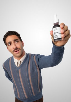 Mężczyzna z wąsami i w paskowanym swetrze trzyma i pokazuje butelkę z kroplomierzem oznaczoną „The Ordinary” na jednolitym tle.