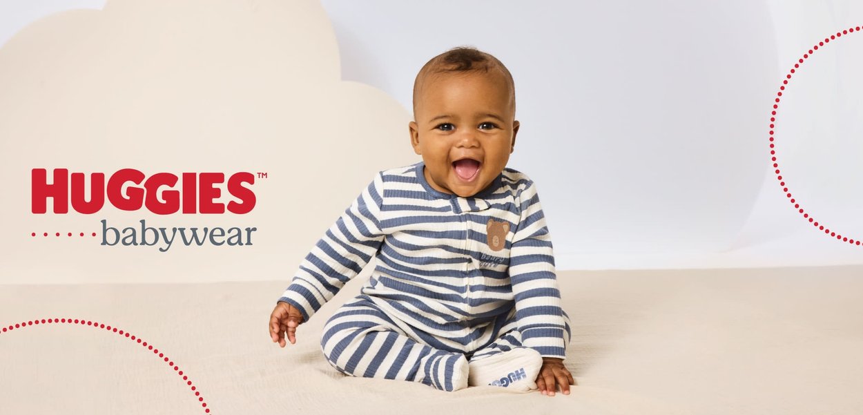 Bambino sorridente in pigiama a righe blu e bianche seduto su una superficie beige con il logo Huggies babywear sopra.
