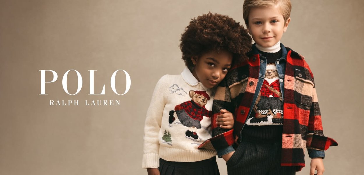 Due bambini che indossano maglioni festivi e abbigliamento a strati stanno vicini, uno appoggiato sulla spalla dell'altro contro uno sfondo semplice.