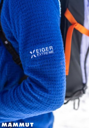 Manche en polaire texturée bleue avec le logo "Eiger Extreme", faisant partie de l'équipement de plein air porté par une personne portant un sac à dos noir et orange.