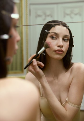 Eine Person trägt mit einem Make-up-Pinsel Rouge auf, steht vor einem Spiegel und trägt ein hautfarbendes, enganliegendes Kleidungsstück; das Ganze wird von sanfter Beleuchtung und Haarklammern umrahmt.