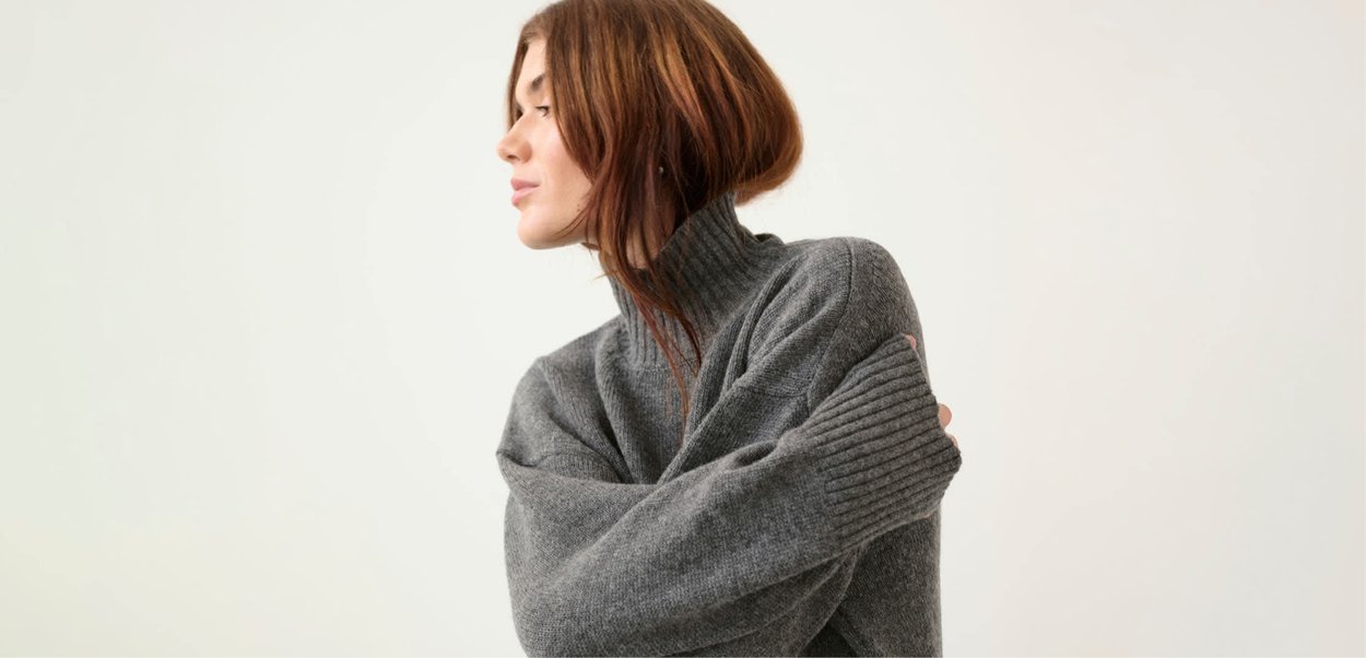 Harmaa ylipitkä neulepaita, jossa korkea ribbineulekaulus ja hihansuissa ribbineuletta. Pehmeä koostumus. Malli kääntyy sivuttain.