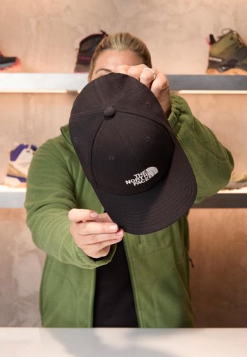 Schwarze Baseballkappe mit gebogenem Schirm und gesticktem weißen Logo von "The North Face", die von einer Person in einer grünen Jacke gehalten wird.