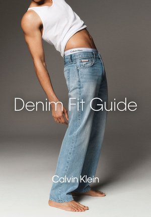 Mand læner sig tilbage barfodet iført lysblå vide jeans og en hvid tank top, der viser Calvin Klein's talje og logo.