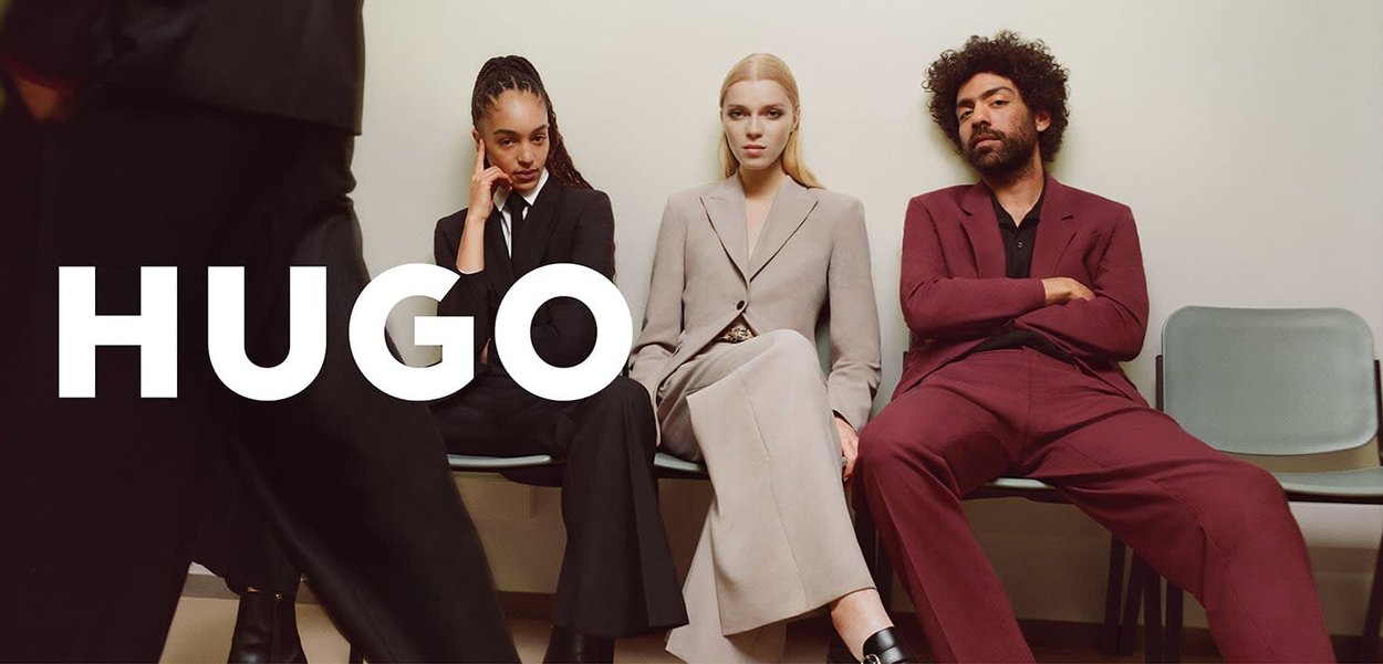 Trois personnes sont assises sur des chaises portant des costumes sur mesure : gris clair, noir et bordeaux. L'espace présente un arrière-plan neutre et une décoration minimaliste.