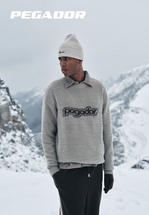 Grau melierter Pullover mit "Pegador"-Logo, mit Kragen, über einem langärmeligen Hemd getragen; kombiniert mit dunklen Hosen und einer hellgrauen Mütze.