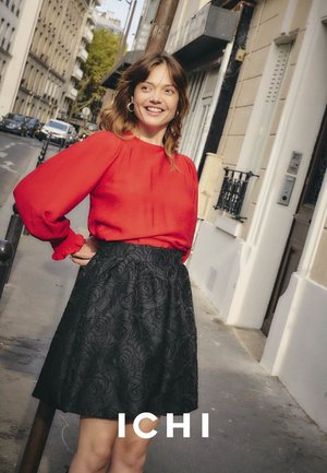 Rote Bluse mit langen Ärmeln und gerafften Manschetten, kombiniert mit einem strukturierten schwarzen Rock mit Rosenmuster. Urbane Kulisse mit Straßenelementen.