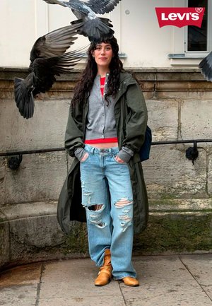 Une femme se tient contre un mur en pierre, portant un jean bleu déchiré, un long manteau vert, un cardigan gris et des bottes couleur fauve ; des pigeons volent au-dessus ; logo Levi’s en haut à droite.