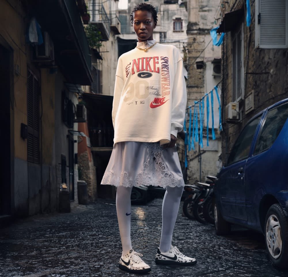 Felpa bianca Nike oversize con loghi rossi e neri, indossata sopra una gonna grigia chiara con bordi in pizzo, abbinata a sneaker con un motivo in bianco e nero.