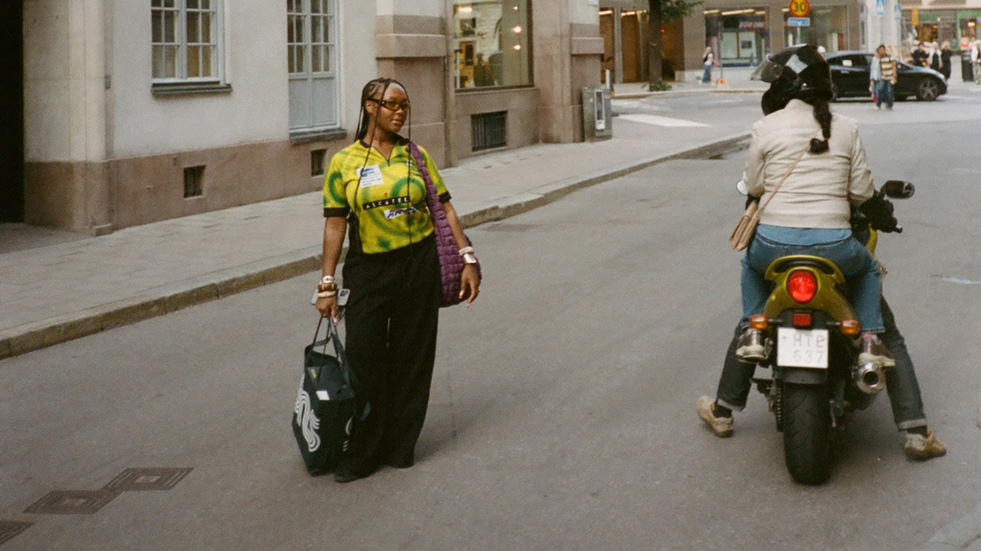 Een persoon in een groen en geel geblokt shirt en zwarte broek loopt met een grote donkere tas, terwijl er een motorfiets in de buurt passeert.