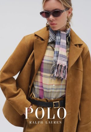 Mujer con chaqueta y pantalones de ante marrón, camisa de cuadros, bufanda de cuadros multicolor, grandes pendientes de aro y gafas de sol oscuras.