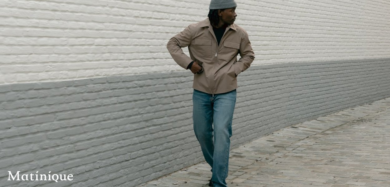 Mann trägt eine beige Jacke, blaue Jeans und eine graue Mütze, geht entlang einer zweifarbigen Backsteinmauer auf einem gepflasterten Weg.