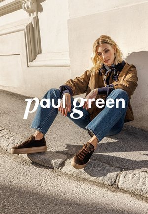 Brown suede sneakers with a beige rubber sole, worn with blue jeans and a brown jacket, set against a light-colored wall.