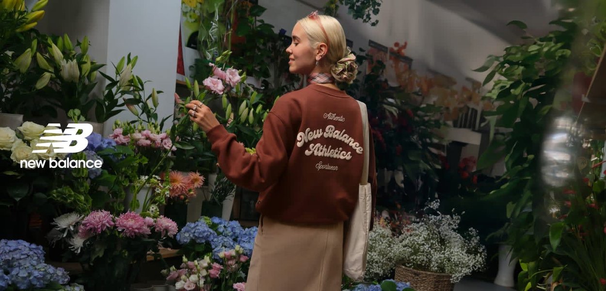 Bruine sweatshirt met de tekst "New Balance Athletics" in crèmekleur, gecombineerd met een beige rok. Omringd door een verscheidenheid aan kleurrijke bloemen en groen.