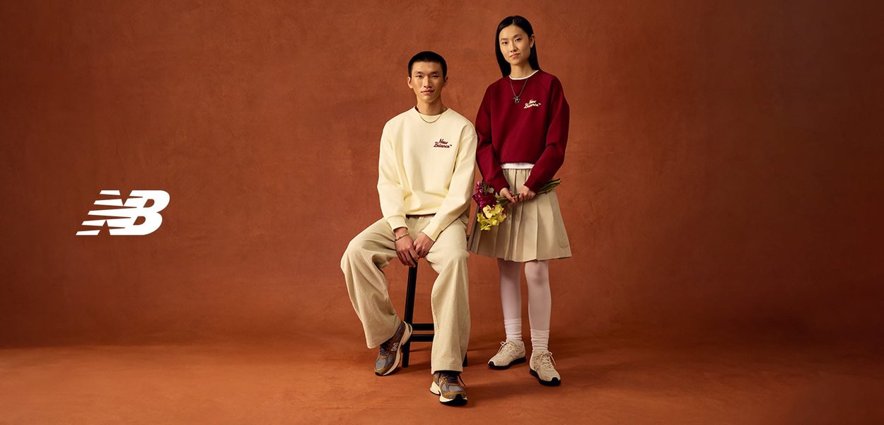 Deux mannequins posent devant un fond marron ; l'un est assis, portant un sweat-shirt crème et un pantalon beige, l'autre est debout, vêtu d'un sweat-shirt rouge et d'une jupe beige, tenant des fleurs.