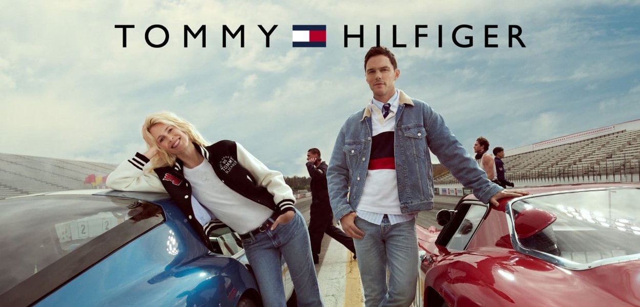 Ein blauer Sportwagen und ein rotes Vintage-Auto in einer Rennstrecken-Umgebung. Zwei Personen in lässiger Kleidung, darunter eine schwarz-weiße Jacke, Jeans und ein gestreiftes Oberteil.