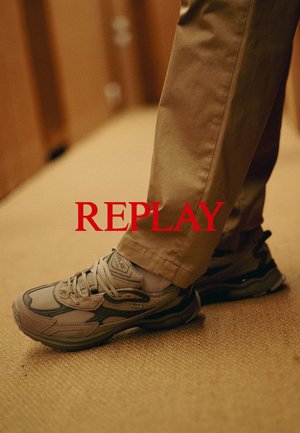 Lichtgrijze sport sneakers met donkere accenten, gaas en textuur overlays, en een dikke zool. Draagt met beige katoenen broeken op een gestructureerd oppervlak.