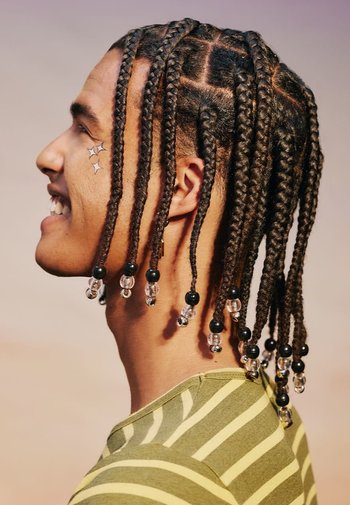 Des cheveux tressés ornés de perles transparentes et noires, profil latéral d'une personne portant une chemise rayée verte, mettant en valeur un design capillaire complexe.