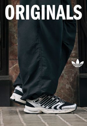 Scarpe sportive in bianco e nero con tomaia in mesh testurizzato, dettagli argento e suola ammortizzata, abbinate a pantaloni scuri.