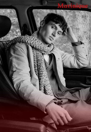 Hellfarbige Jacke mit einem strukturierten, gestrickten Schal, der über die Schulter gelegt ist, sitzend in einem dunklen Autoinnenraum. Sanfte, gedämpfte Farben, lässige Haltung.