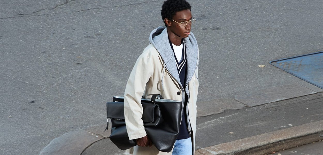 Lichtbeige trenchcoat met een grijze hoodie eronder, blauwe jeans en een grote zwarte handtas. Straatscene met een asfalt achtergrond.