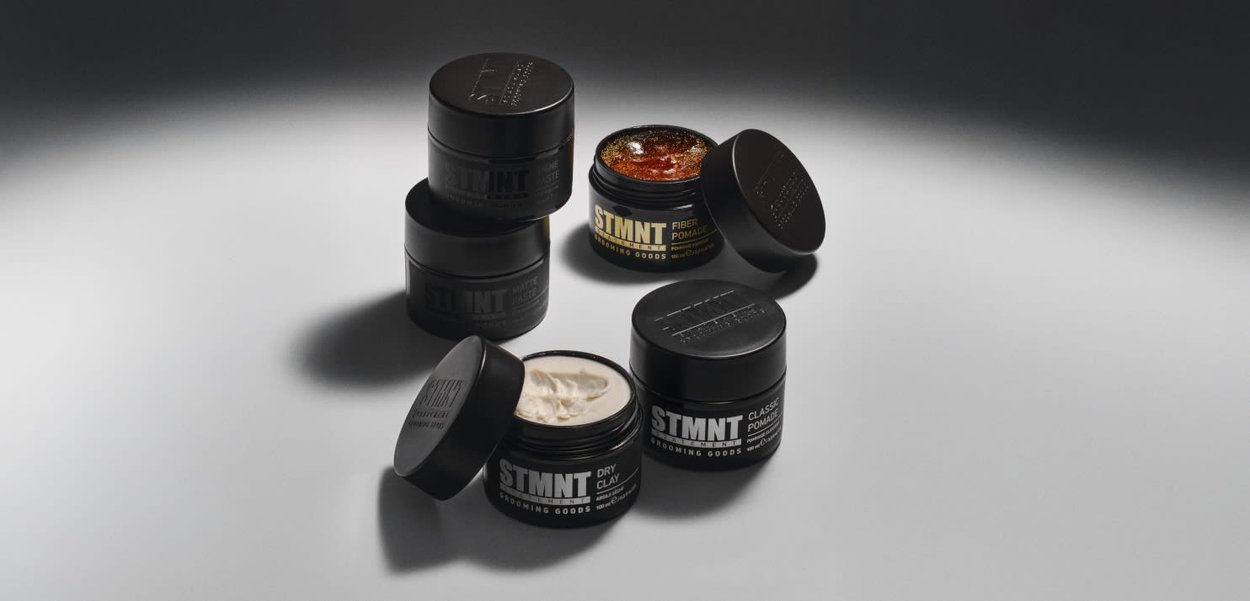 Fünf schwarze zylindrische Dosen mit Haarpflegeprodukten und markanten Etiketten. Deckel sind mit matter Textur und glänzenden Oberflächen versehen; Inhalt variiert in Farbe und Konsistenz.