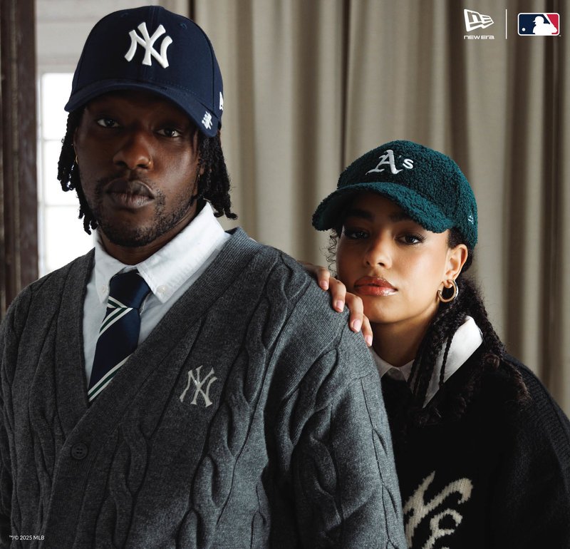 Uomo con cappellino dei Yankees blu navy e cardigan grigio, donna dietro di lui con cappellino verde degli Athletics e maglione nero, entrambi guardano verso la fotocamera.