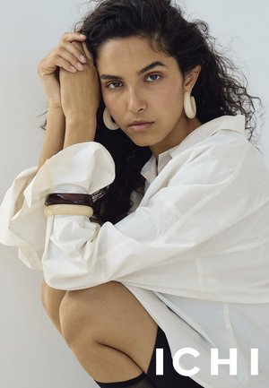 Kvinde med krøllet mørkt hår iført oversized hvid skjorte, store hoop-øreringe og chunky armbånd, mens hun sidder med armene foldet nær ansigtet.