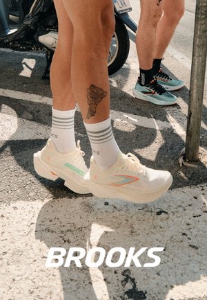 Zapatillas de running blancas con acentos coloridos, parte superior texturizada y suela gruesa. Calcetines a rayas grises y un tatuaje en la pierna, fondo urbano.