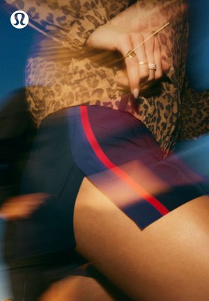 Persona con camiseta de estampado de leopardo y pantalones cortos azul marino con rayas rojas, con manos entrelazadas que llevan anillos y uñas largas, con un ligero efecto de desenfoque por movimiento.